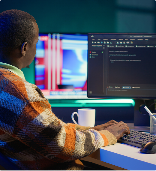 Pessoa digitando código em IDE de tema escuro, sentada à mesa com caneca ao lado, monitores ao fundo.